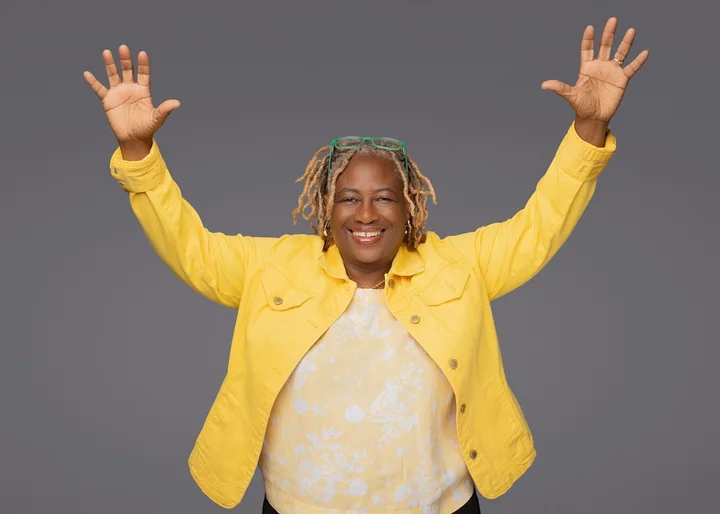 Vena Nelson, CEO of Go Big Accounting, smiling with both arms raised, in a yellow jacket over a yellow floral top, in front of a plain grey backdrop.